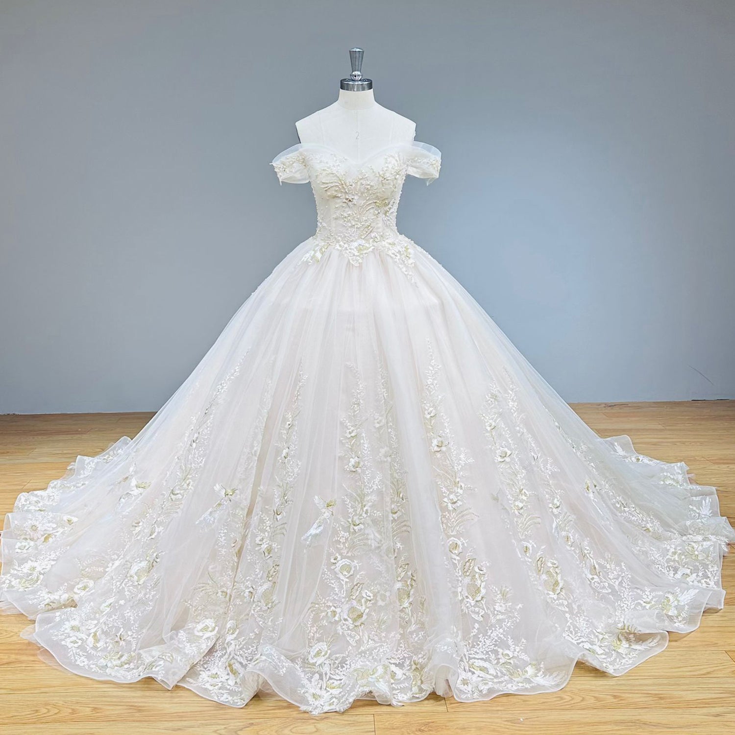 Front display of an off-the-shoulder princess bridal gown featuring detailed trailing floral lace appliques across a voluminous skirt.