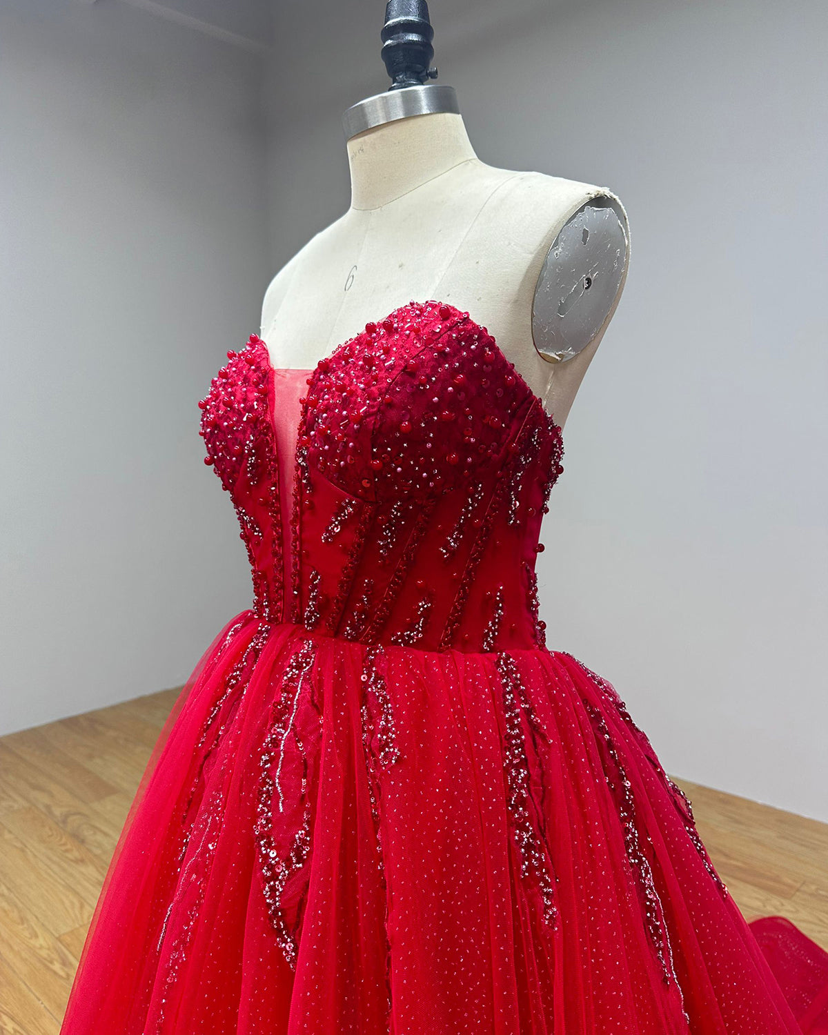 Side profile detail of a red sweetheart neck wedding dress on a dress form, showcasing the structured corset boning, thick hand-beaded crystal embellishments, and layered tulle glitter skirt.