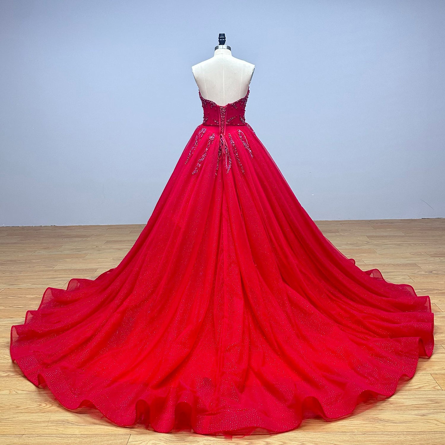 Full back view of a magnificent crimson wedding gown on a dress form, displaying a classic lace-up back and an expansive chapel-length train crafted from shimmering glitter tulle.