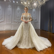 Front view of an elegant bride wearing a luxury off the shoulder straps mermaid wedding dress featuring a voluminous removable overskirt, a plunging illusion neckline, and intricate shimmering crystal beadwork.