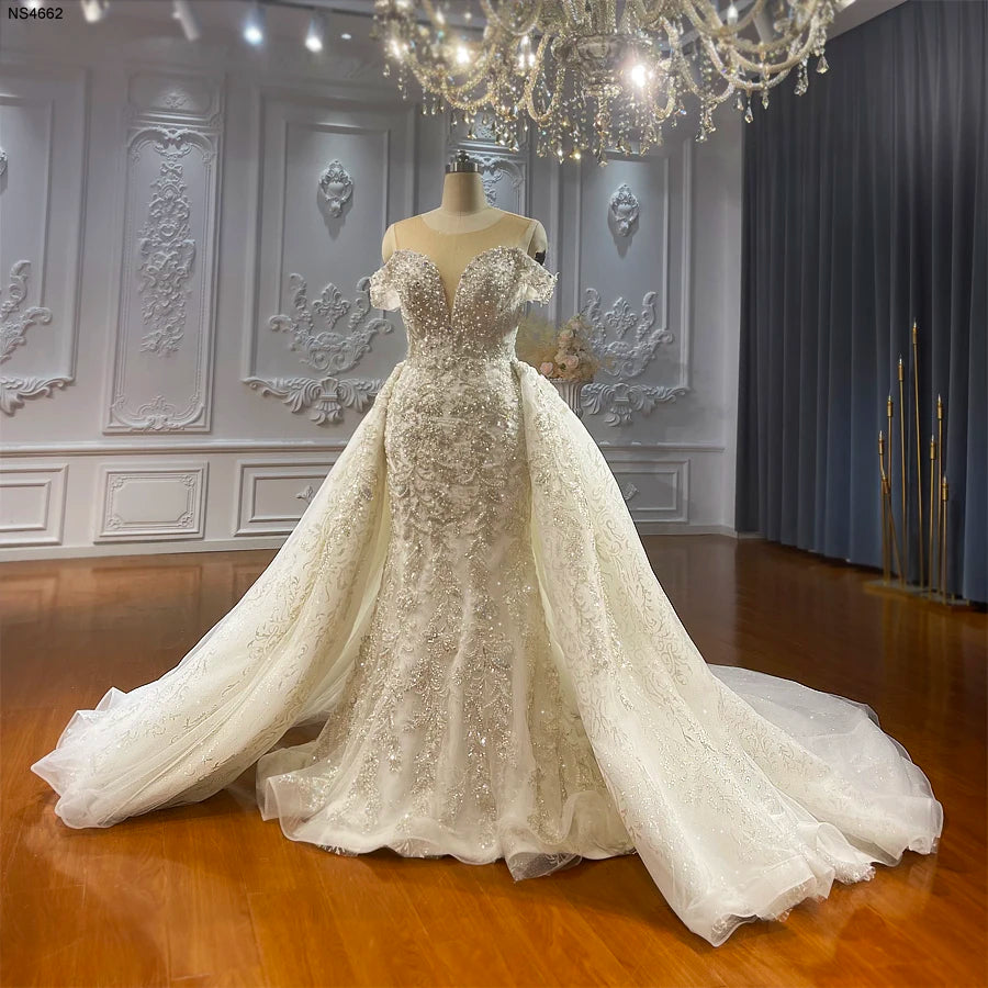 Front view of a luxury off-the-shoulder mermaid wedding dress with a voluminous removable ball gown overskirt, featuring heavy crystal beading and intricate floral lace appliques.