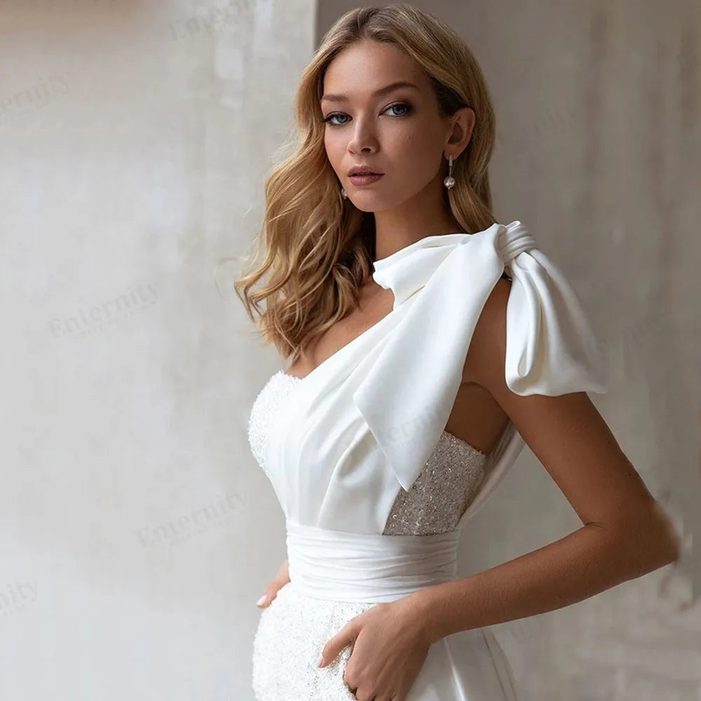 Detailed view of a one-shoulder bridal jumpsuit bodice highlighting an elegant draped white satin neckline overlay with an attached statement shoulder bow.
