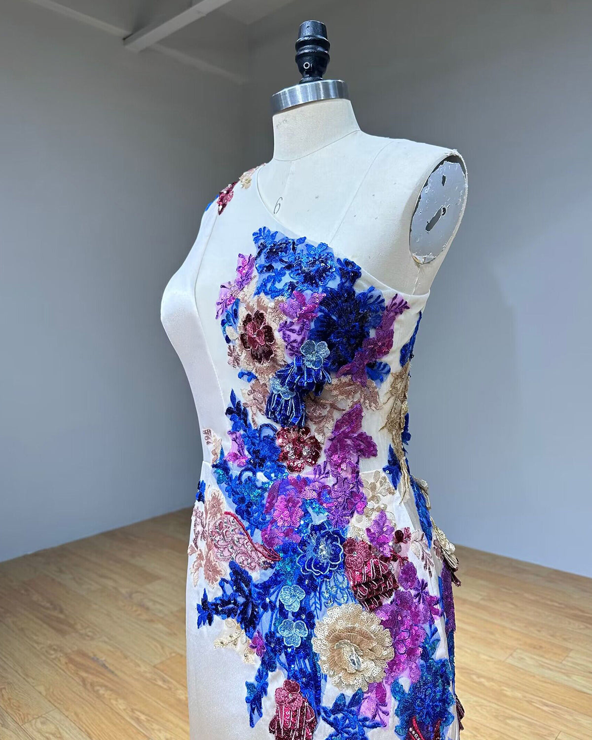 Side profile studio shot focusing on the high leg slit drape and flowing column silhouette of a white one-shoulder formal dress on a mannequin.