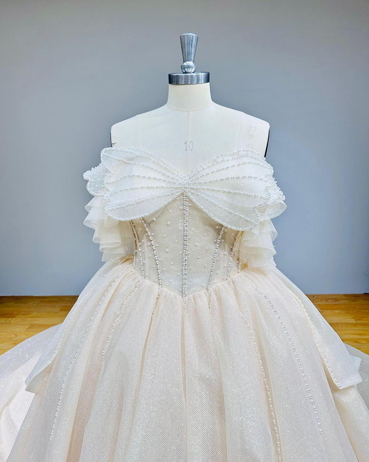 Close-up front bodice detail of a glitter ball gown wedding dress on a mannequin, detailing the unique pearl-embellished bow across the chest, structured corset boning, and off-shoulder ruffled tulle sleeves.