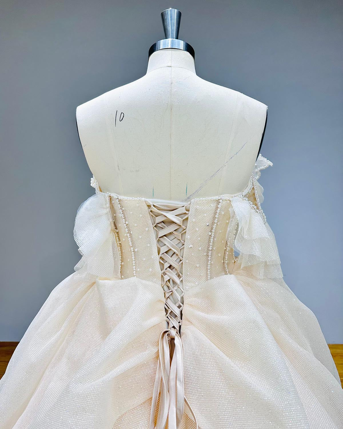 Close-up back bodice view of a pearl bow wedding dress on a mannequin, focusing on the traditional lace-up back corset, pearl-lined seams, and delicate off-shoulder ruffled sleeves.