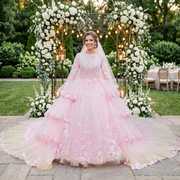 Smiling bride wearing a vibrant pink long-sleeve wedding dress with a dramatic tiered tulle ball gown skirt, standing outdoors under a white floral archway.