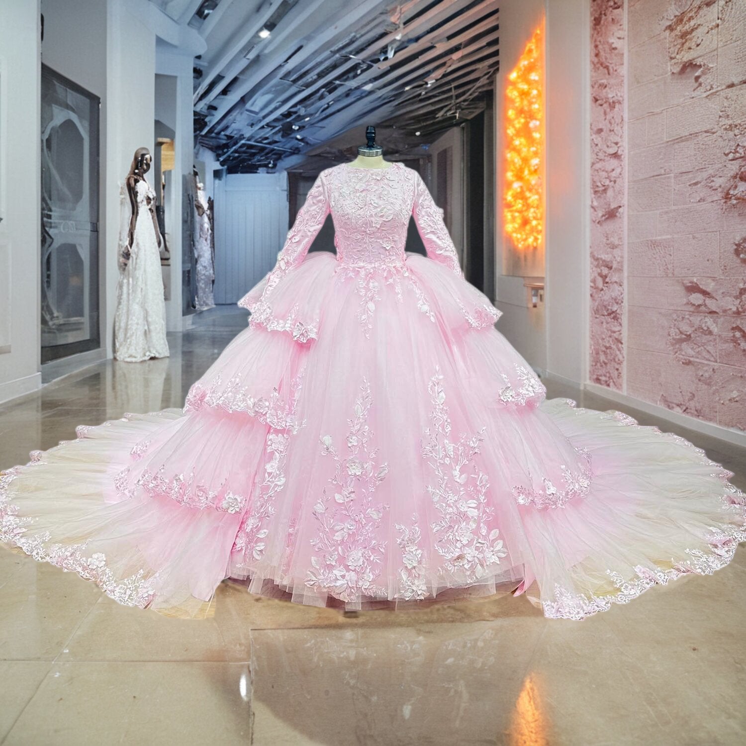 Pink A-line modest wedding gown with long embroidered sleeves and a tiered tulle skirt panel overskirt displayed on a mannequin inside a boutique showroom.