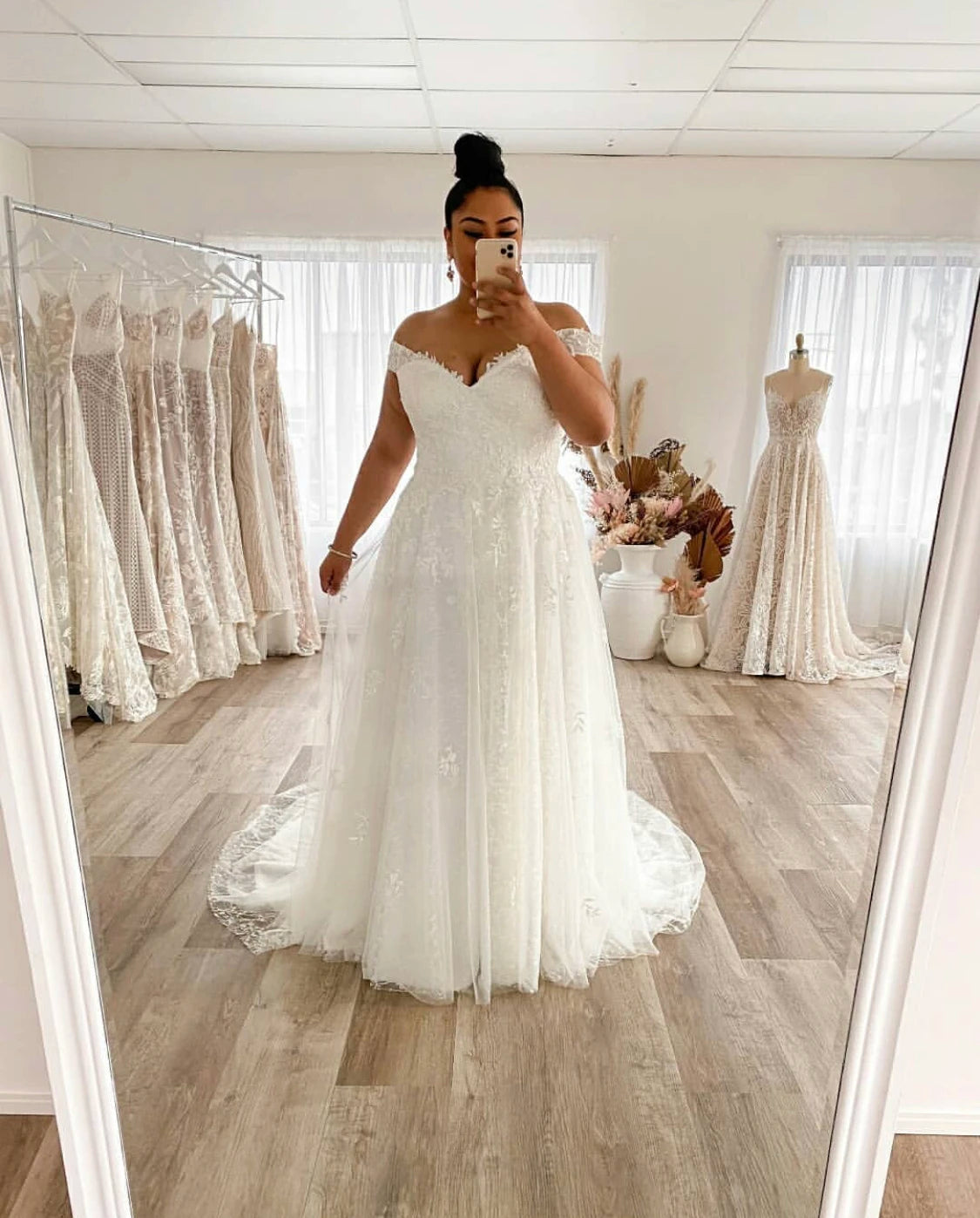 A plus-size bride smiling in a mirror, wearing an off-shoulder A-line wedding dress with delicate lace applique and a flowing tulle skirt.
