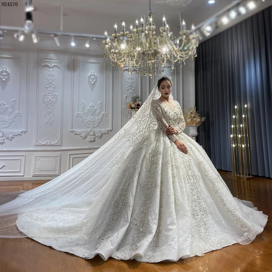 Full-length front view of an elegant model in a luxury plus size ball gown wedding dress featuring shimmering crystal embellishments, long sleeves, and a grand cathedral train with a matching beaded veil.