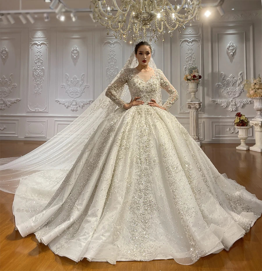Elegant front perspective of a long sleeve ball gown bridal gown showcasing the intricate hand-beaded lace bodice, a soft V-neckline, and an expansive sparkly skirt in a luxury bridal showroom.