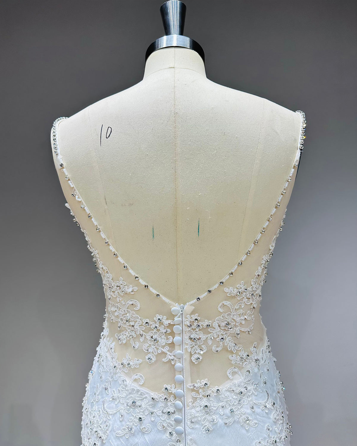 Close-up of the back of a bridal gown highlighting a low illusion V-back, delicate crystal-trimmed straps, and fabric-covered buttons over a zipper closure.