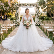 A smiling bride wearing a sparkling sequined ball gown wedding dress with a plunge V-neckline and sheer tulle cape sleeves, holding a bridal bouquet at an outdoor garden wedding altar.