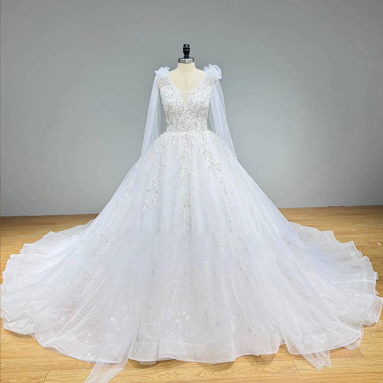 A front view of a sequined and beaded ball gown bridal gown on a dress form, highlighting the sheer shoulder bows, flowing cape elements, and a wide A-line tulle skirt.