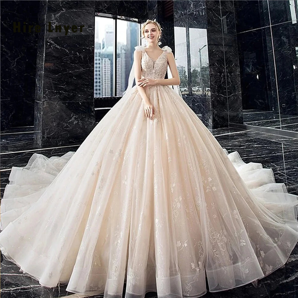 A blonde model wearing a champagne-tinted royal ball gown wedding dress with a glistening crystal bodice and a full dramatic tulle skirt, posing in a modern luxury indoor setting.