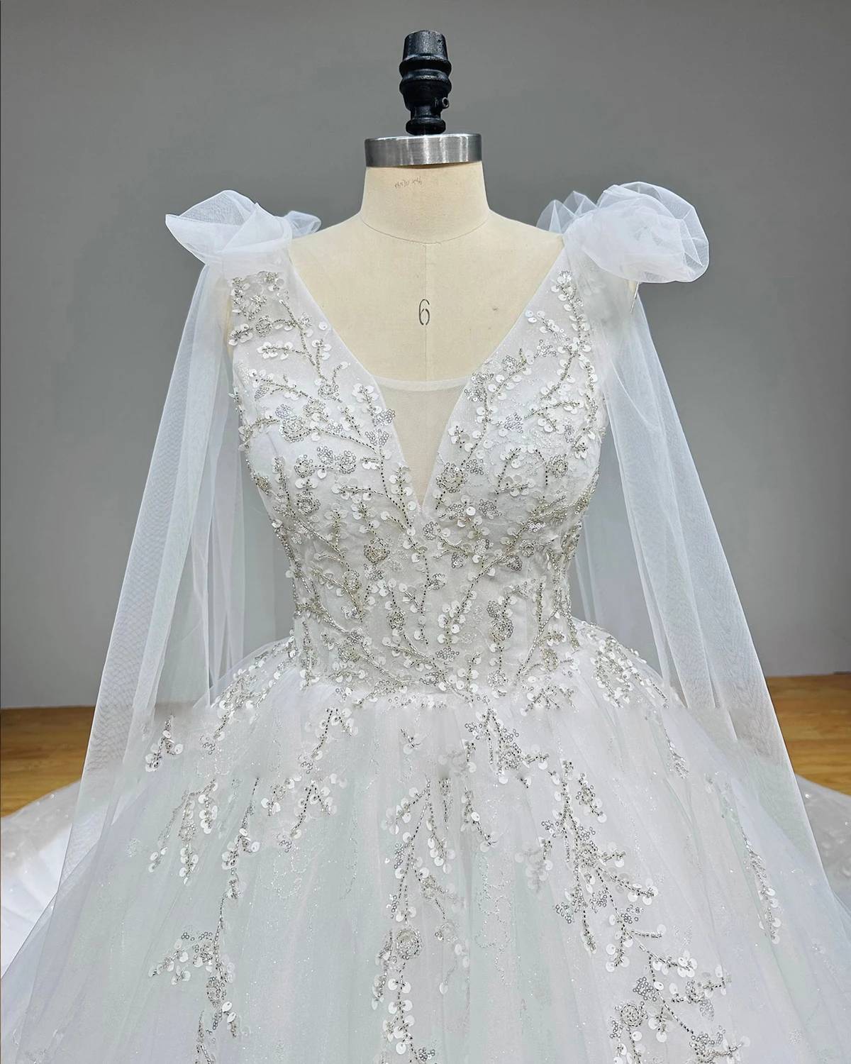 Close-up bodice view of a luxury wedding dress featuring a sheer illusion plunge V-neckline decorated with intricate floral sequin patterns and metallic beadwork.