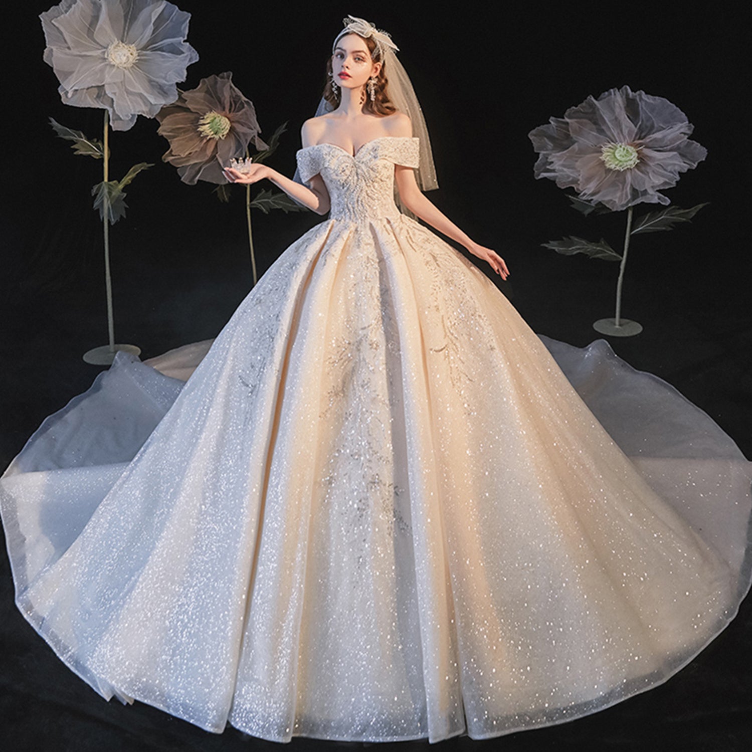 Editorial bridal model posing in a shimmering champagne off-the-shoulder princess ball gown wedding dress with sequin appliques in a dramatic dark studio setting.