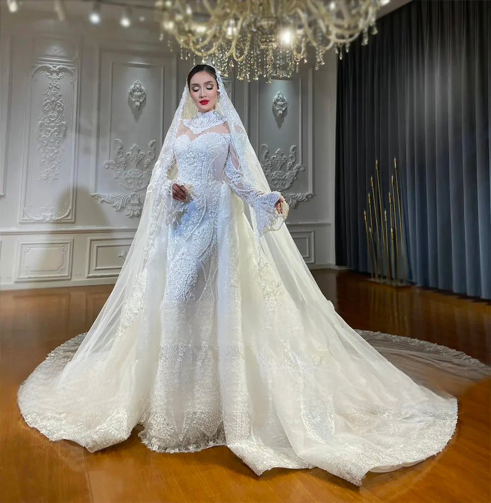 A radiant bride modeling the versatile 2-in-1 lace mermaid wedding dress with the dramatic removable overskirt attached, featuring sheer long sleeves and an elegant bridal veil in a luxury showroom.