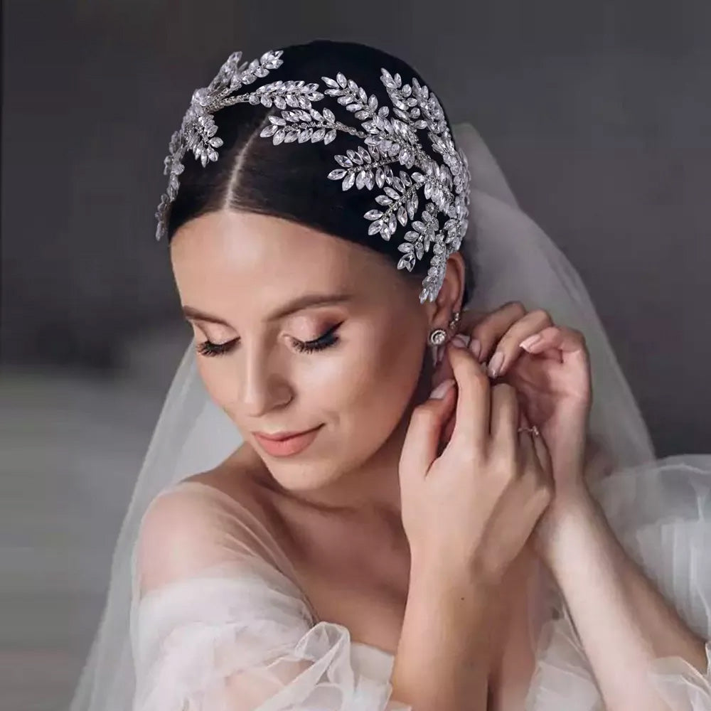 Close-up portrait of a bride adjusting her earring, highlighting a sleek silver rhinestone wedding headband structured with rows of crystal leaf branches.