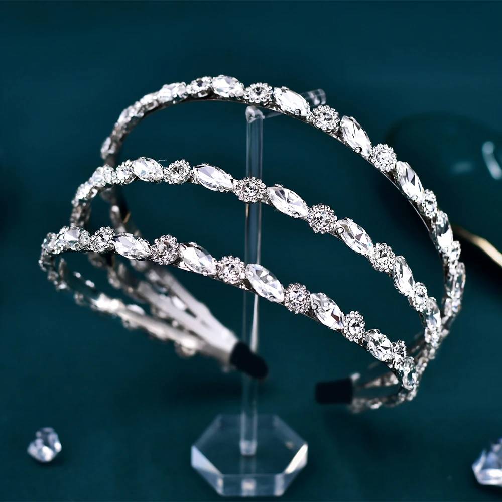 A three-row silver crystal wedding tiara and headband displayed on a clear stand against a dark teal background.