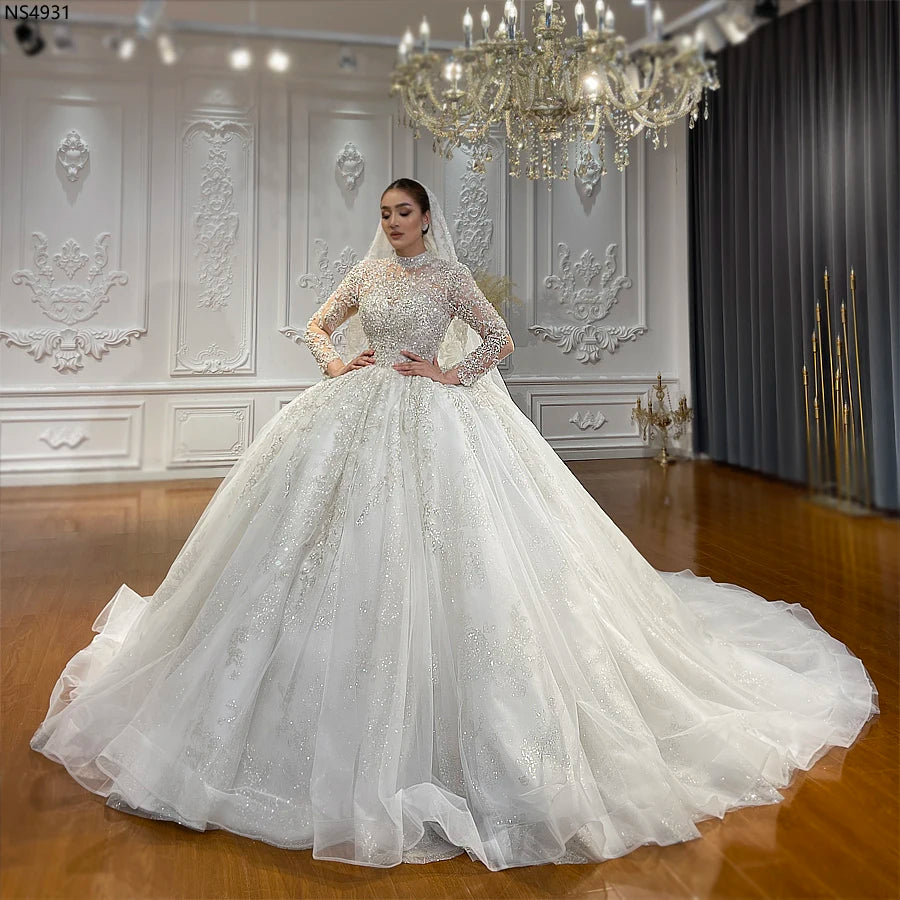 Full view of a romantic high neck lace wedding gown featuring a grand, sparkling ball gown skirt and elegant long sleeves, modeled in a bright bridal boutique.