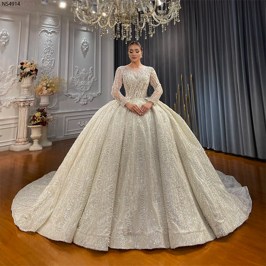 Full front view of a bride wearing a romantic long sleeve ball gown wedding dress featuring intricate pearl detailing, a high illusion neckline, and a voluminous sparkling skirt.