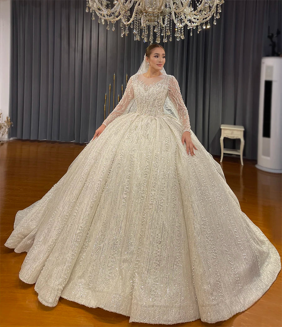 Front view of a glamorous bride in a luxury pearl-embellished long sleeve wedding gown with an illusion neckline, posing elegantly in a bridal showroom.