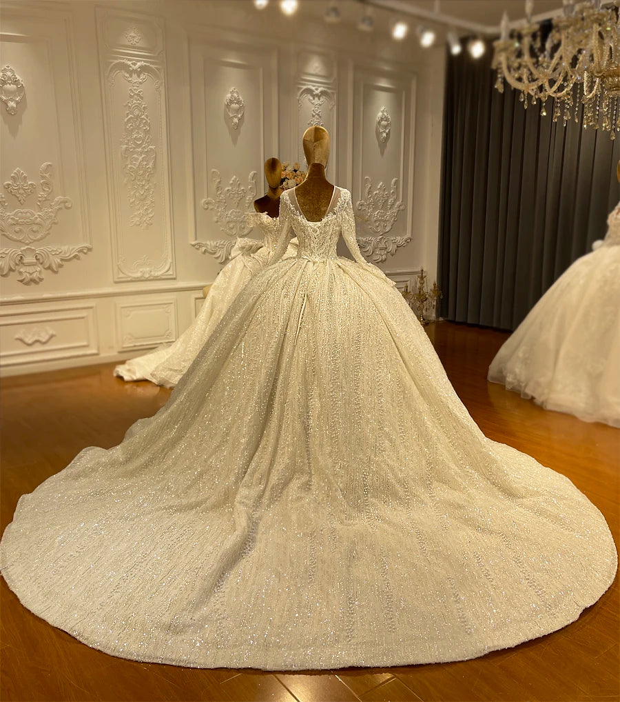 Rear view of a romantic long sleeve ball gown wedding dress on a mannequin, highlighting the elegant keyhole back, lace-up corset, and long shimmering cathedral train covered in pearls.