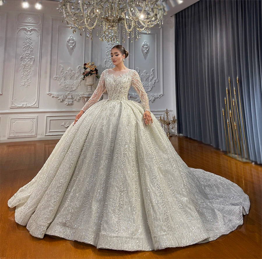 Full front view of a model showcasing a romantic long sleeve ball gown wedding dress with heavy pearl detailing and a grand, flared silhouette under a chandelier.