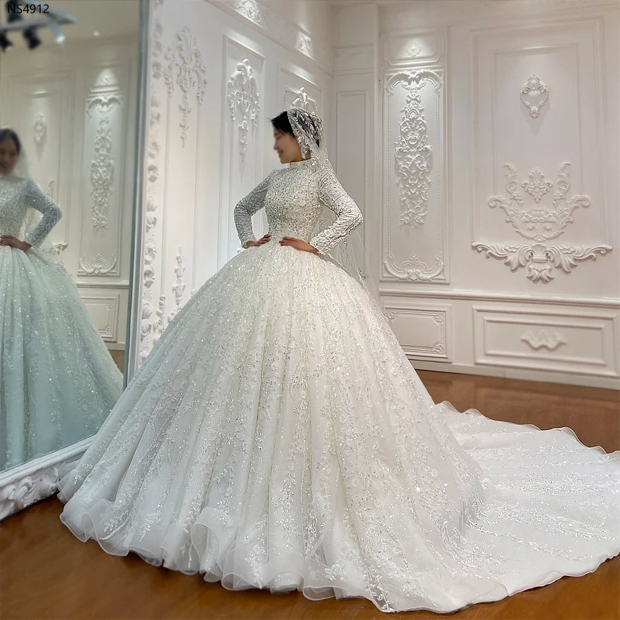 Front-angle view of a bride modeling a royal Islamic wedding dress with a modest high neckline, intricate handmade embroidery, and a matching embellished hijab in a luxury bridal suite.