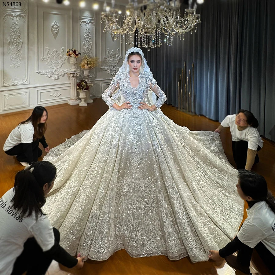 A bride in a heavy beaded royal V-neck luxury ball gown wedding dress with long sleeves, being assisted by a professional team to showcase the expansive shimmering cathedral train.
