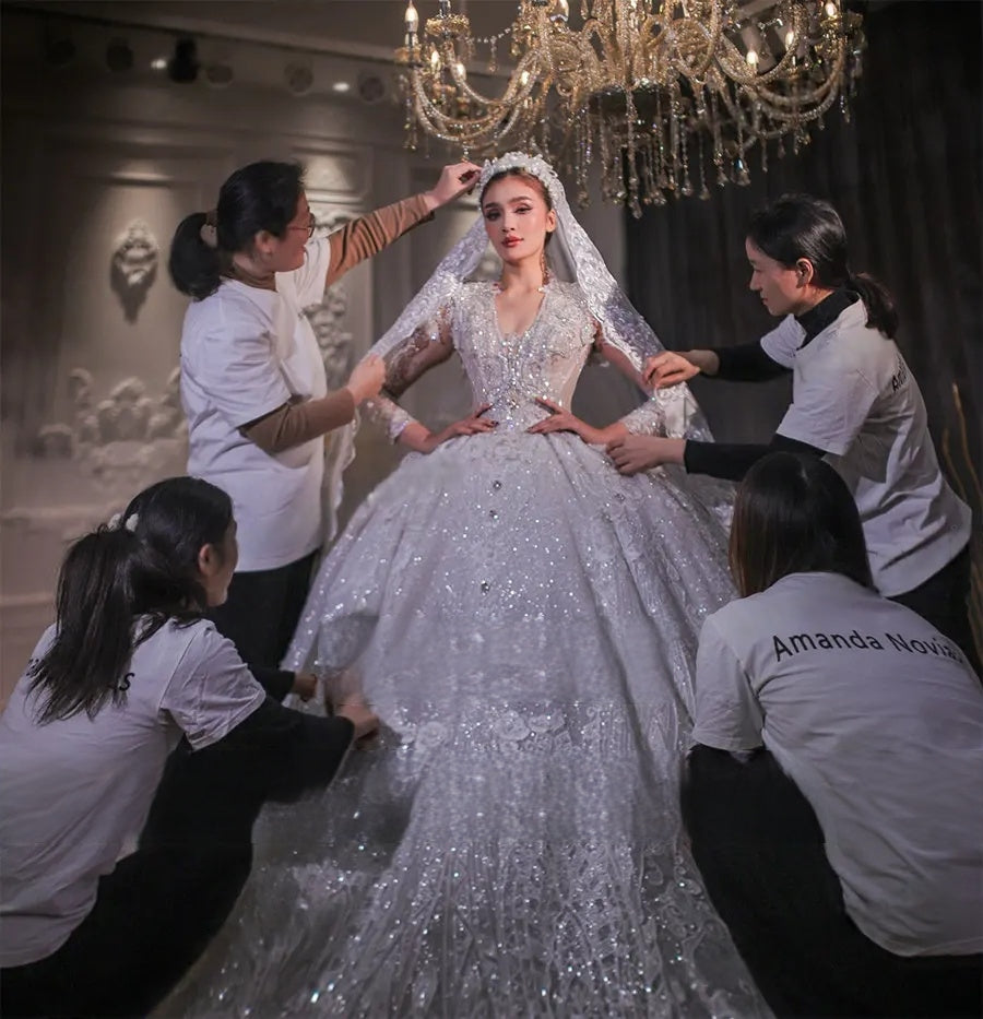 Close-up shot of a royal V-neck bridal gown bodice highlighting the intricate silver beadwork and sheer long sleeves being adjusted under a crystal chandelier.