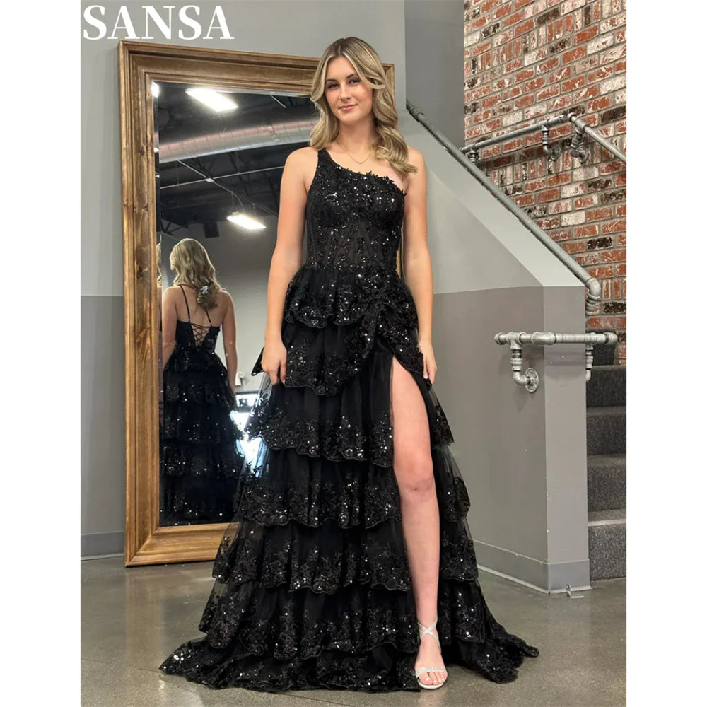 Model wearing a black Sansa glitter one-shoulder A-line evening dress with a sheer floral lace bodice, tiered floor-length ruffled skirt, and high leg slit.