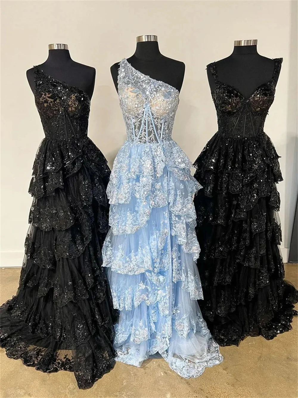 Three mannequins displaying the Sansa glitter evening dress in black one-shoulder, light blue one-shoulder, and black sleeveless styles with tiered multilayer lace skirts.