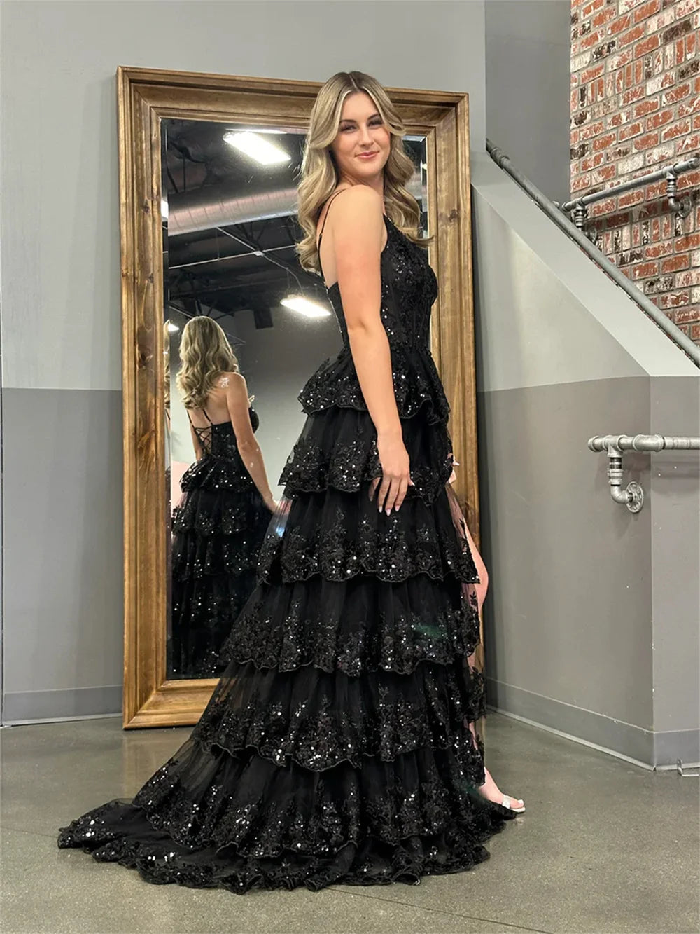 Side view of the black Sansa glitter tiered evening dress showcasing the volume of the multilayer floor-length skirt and the corset-style lace-up back detail in the mirror reflection.