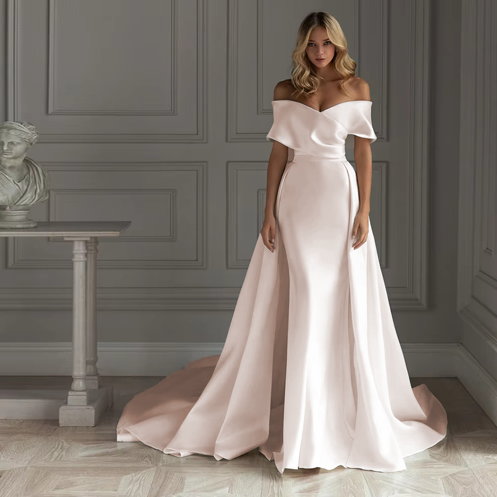 Romantic soft blush pink satin mermaid wedding dress featuring an architectural off-the-shoulder portrait sleeve cuff and an attached matching pink overskirt train.