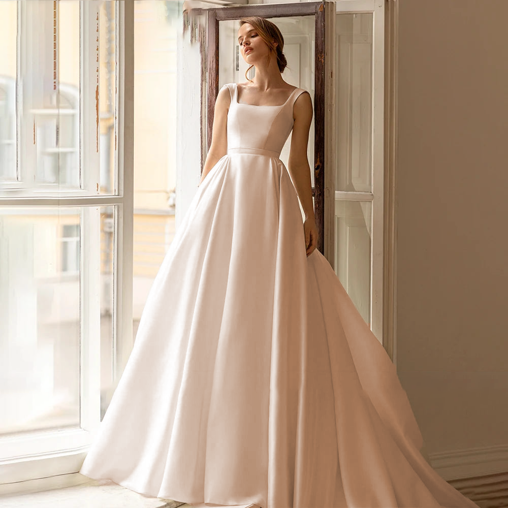 A bride standing by a window wearing a classic, sleeveless ivory A-line wedding gown with a clean and minimal satin finish.