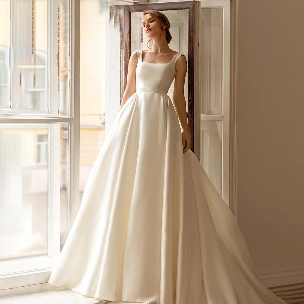 A full-length view of a bride modeling a sleeveless ivory A-line wedding dress by a window, highlighting the graceful silhouette.
