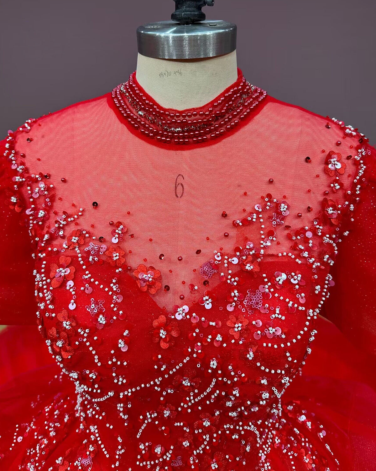 Intricate close-up of the front bodice of a red bridal gown, highlighting the sheer illusion high neck, beaded collar, sequins, and 3D floral appliques.
