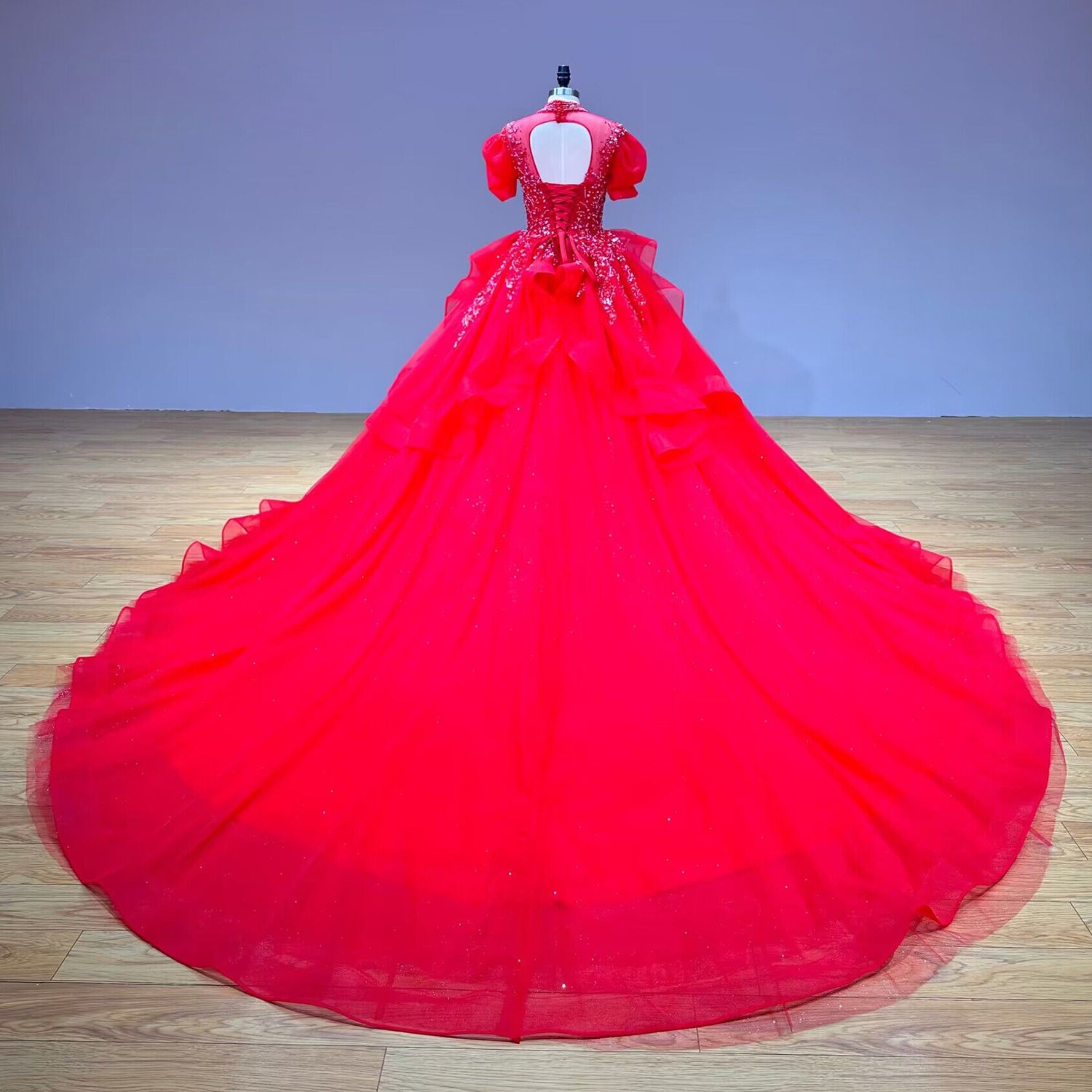 Full back display of a red sequin ball gown wedding dress on a mannequin, highlighting the keyhole back, corset tie, and an expansive, sweeping circular tulle train.