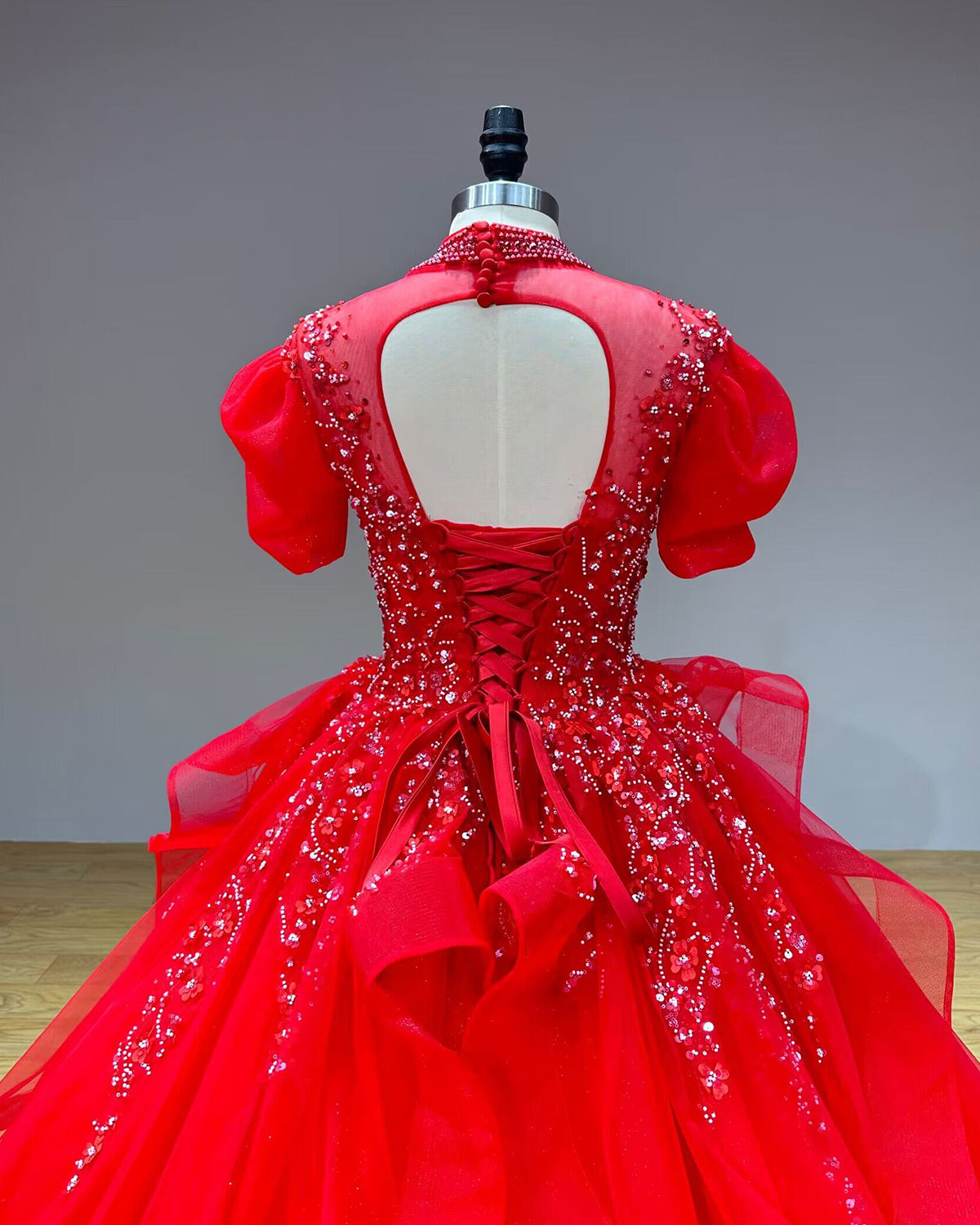 Detailed back view of a red ball gown on a mannequin, showcasing the sheer keyhole back design, adjustable lace-up corset closure, and short puff sleeves.