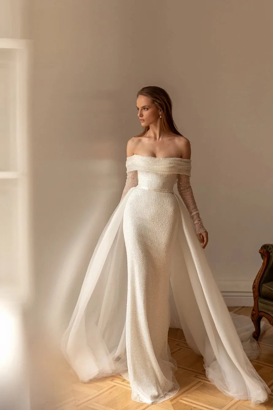 A model showcasing the form-fitting silhouette of an off-shoulder sequin mermaid bridal gown with a sophisticated detachable train.