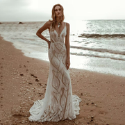 Full-length front view of a model posing on a sandy beach wearing a sexy lace V-neck beach mermaid wedding dress with delicate spaghetti straps and a unique geometric bohemian pattern.