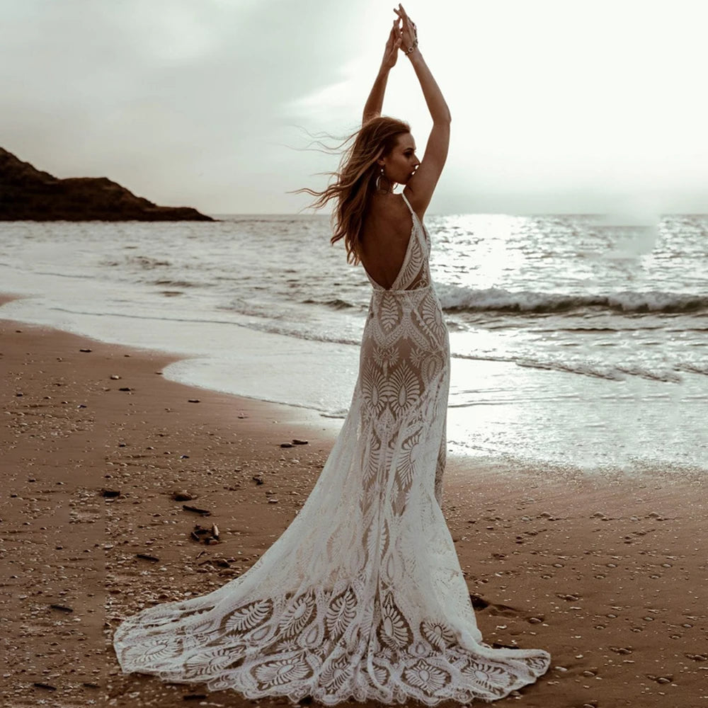 Back view of a bride with arms raised by the shoreline, highlighting the deep backless cut and elegant scalloped lace train of the beach mermaid wedding gown.