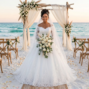 A bride in an elegant long-sleeve, off-shoulder lace wedding gown standing on a sandy beach at sunset, holding a bouquet of white flowers.