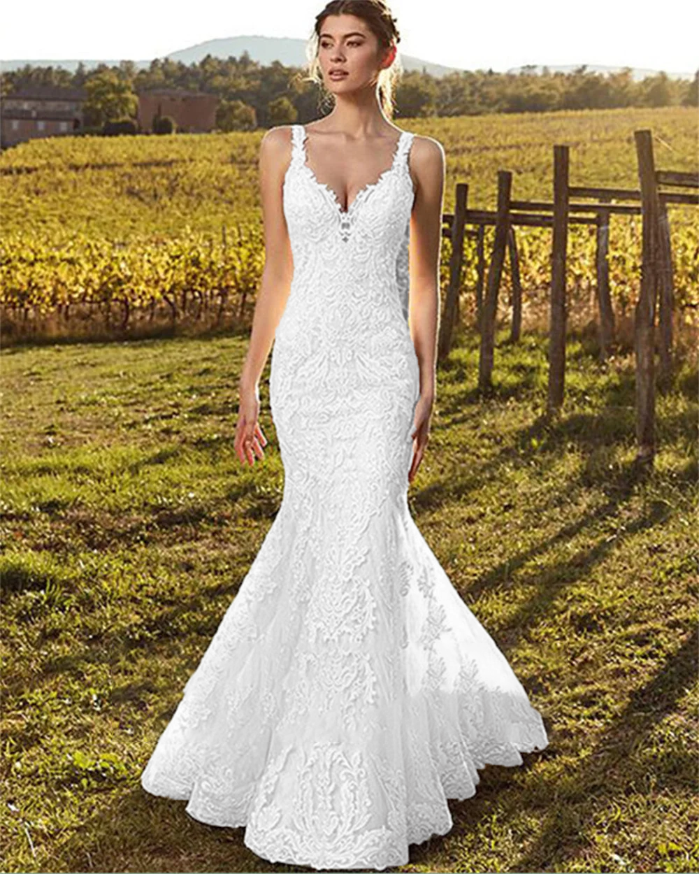A bride standing outdoors in a vineyard wearing a sleeveless, lace-appliqued mermaid wedding gown with a classic V-neck silhouette.