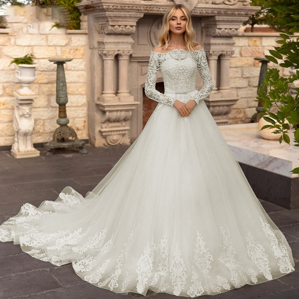 Front view of a model in a luxury A-line bridal gown with long lace sleeves and off-the-shoulder detailing, captured in an elegant architectural setting.