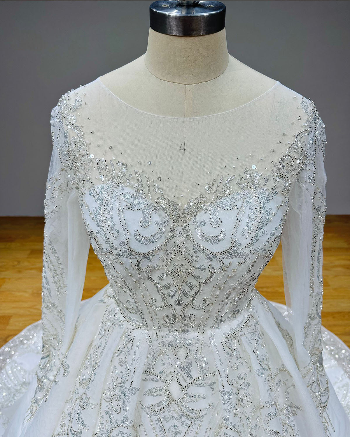 Detailed front close-up of a bridal bodice on a mannequin, showcasing dense silver sequined embroidery, crystal beadwork, and a sheer scoop illusion neckline.