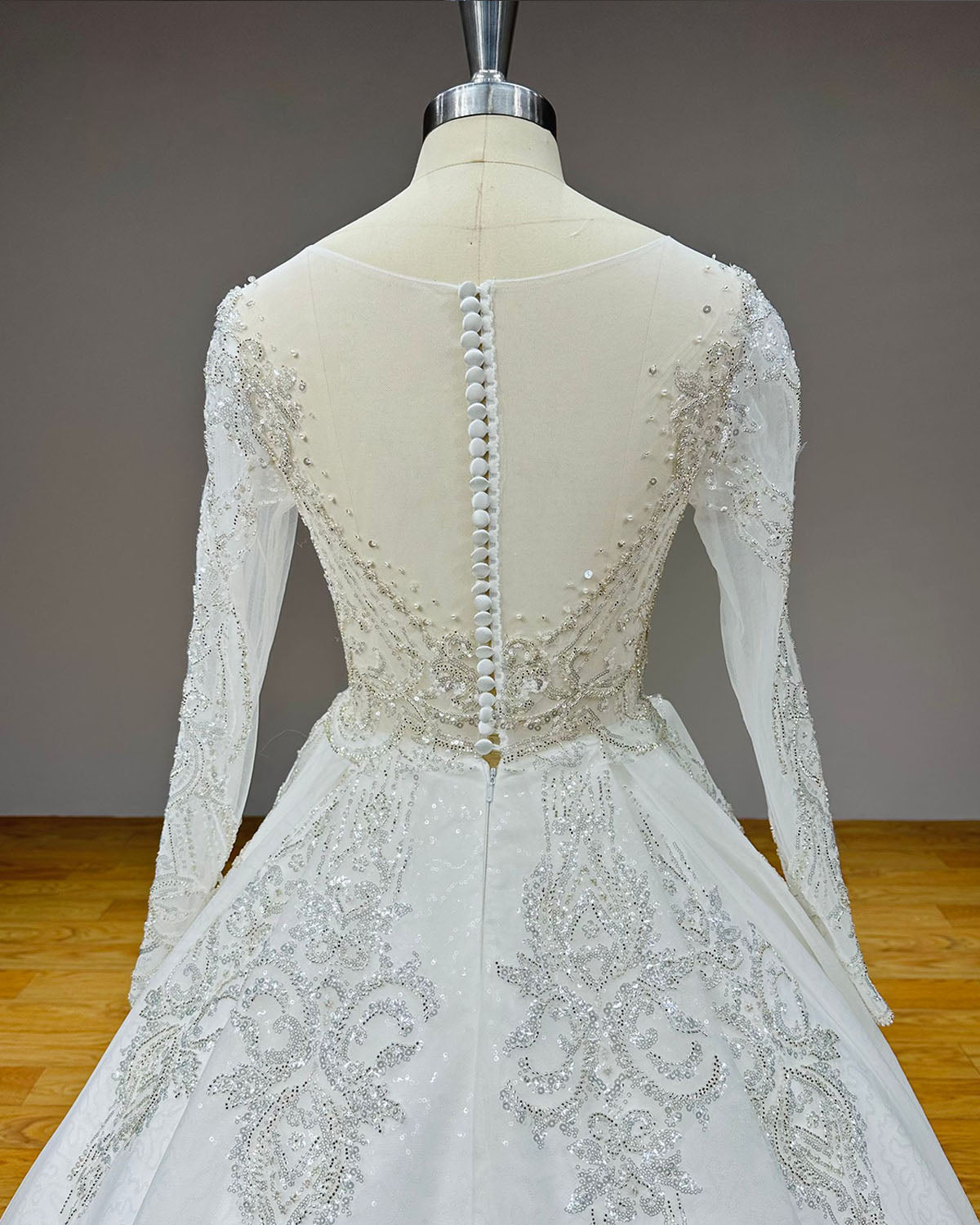Close-up back view of a wedding dress bodice highlighting a sheer illusion back design secured with a long row of elegant fabric-covered buttons and silver sequin scrollwork.