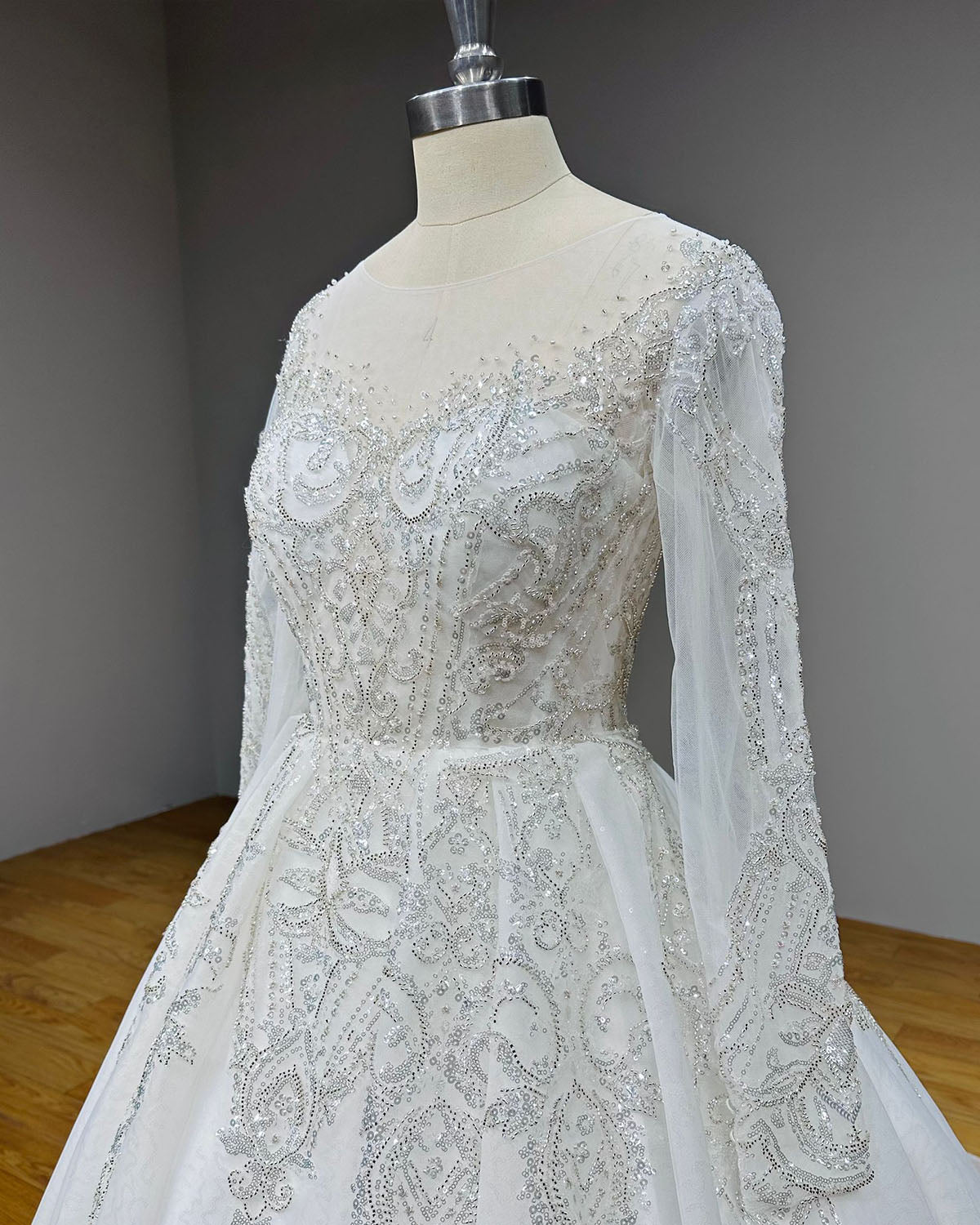 Angled close-up of a sparkling bridal bodice on a dress form, emphasizing the intricate silver sequin patterns along the sheer long sleeves and side panels.