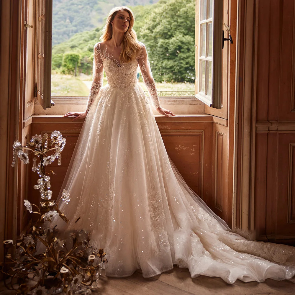 Shining long sleeve wedding dress with a sheer illusion neckline, beaded lace bodice, and a voluminous glitter tulle A-line skirt with a cascading train by a window.